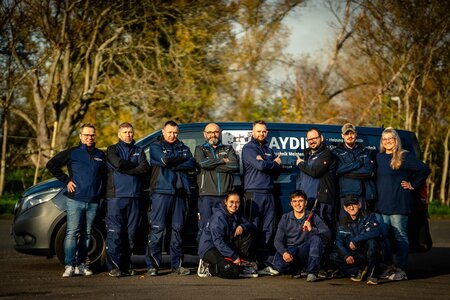 Das Team der Firma Aydin steht draußen vor einem blauen Transporter mit Firmenlogo. Es sind elf Mitarbeiter*Innen zu sehen und sie tragen blaue Firmenkleidung. Einige knien in vorderer Reihe. Im Hintergrund sind herbstliche Bäume zu sehen.