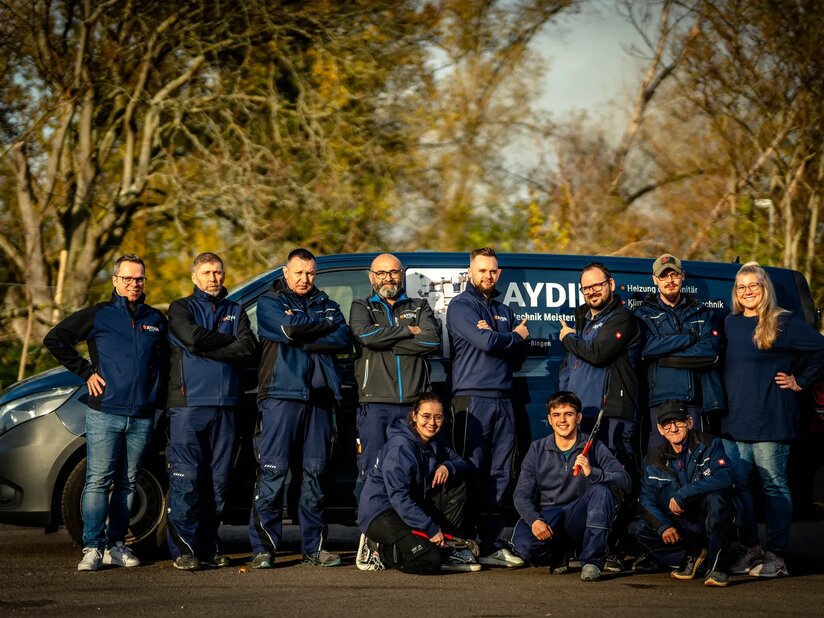 Das Team der Firma Aydin steht draußen vor einem blauen Transporter mit Firmenlogo. Es sind elf Mitarbeiter*Innen zu sehen und sie tragen blaue Firmenkleidung. Einige knien in vorderer Reihe. Im Hintergrund sind herbstliche Bäume zu sehen.