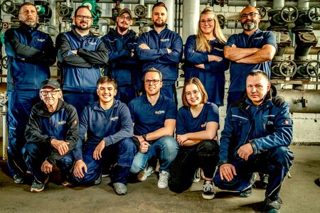 Das Team der Firma Aydin in einer industriellen Umgebung vor technischen Anlagen und Rohrsystemen. Auf dem Bild sind 11 Personen zu sehen.