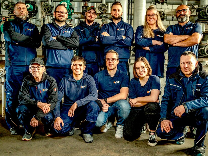 Das Team der Firma Aydin in einer industriellen Umgebung vor technischen Anlagen und Rohrsystemen. Auf dem Bild sind 11 Personen zu sehen.