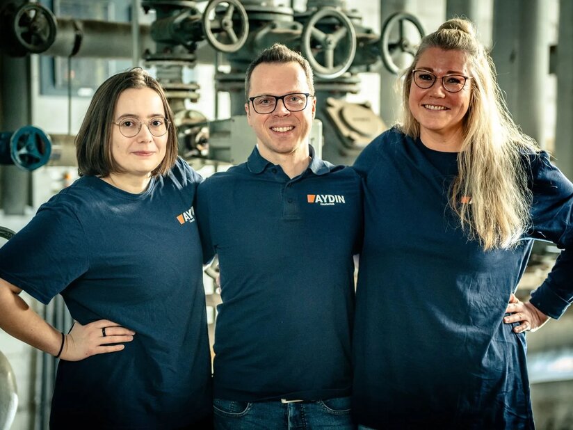 Drei Büro-Mitarbeiter*Innen der Firma Aydin stehen vor einer technischen Anlage und lachen in die Kamera. Sie legen die Arme umeinander, die Mitarbeiter am Rand stemmen eine Hand in die Hüften.