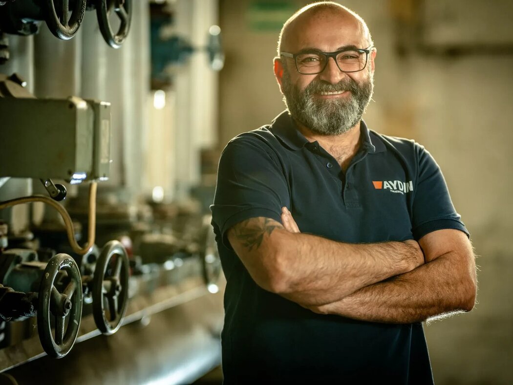 Der Unternehmensleiter Bülent Aydin steht vor einer technischen Anlage mit Ventilrädern. Er trägt ein blaues T-Shirt mit dem Firmenlogo, lacht und hat dabei die Arme verschränkt.