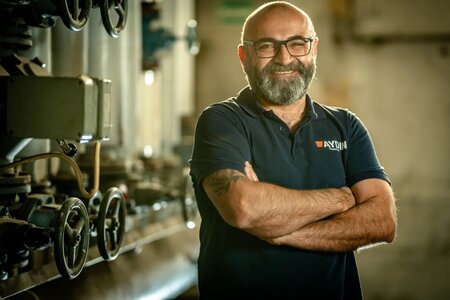 Der Unternehmensleiter Bülent Aydin steht vor einer technischen Anlage mit Ventilrädern. Er trägt ein blaues T-Shirt mit dem Firmenlogo, lacht und hat dabei die Arme verschränkt.