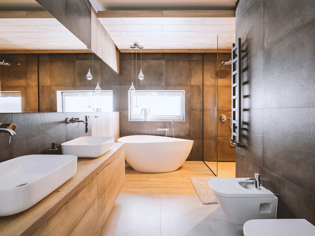 Ein großes modernes Badezimmer mit hölzernem Boden und anthrazit gefliesten Wänden. Hinten im Raum steht eine freistehende Badewanne vor einem Fenster. Links hängt ein breiter hölzerner Waschtisch mit zwei Waschbecken. Rechts befinden sich ein WC, ein Bidet und eine schwarze Handtuchheizung. Eine gläserne Trennwand führt zur Dusche. Stock-Foto.