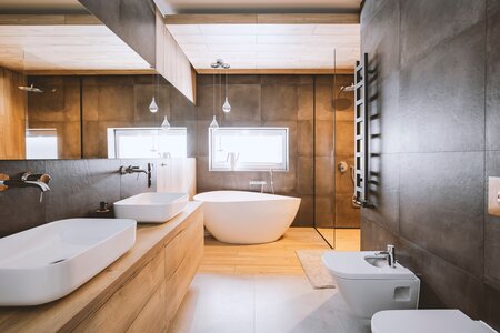 Ein großes modernes Badezimmer mit hölzernem Boden und anthrazit gefliesten Wänden. Hinten im Raum steht eine freistehende Badewanne vor einem Fenster. Links hängt ein breiter hölzerner Waschtisch mit zwei Waschbecken. Rechts befinden sich ein WC, ein Bidet und eine schwarze Handtuchheizung. Eine gläserne Trennwand führt zur Dusche. Stock-Foto.