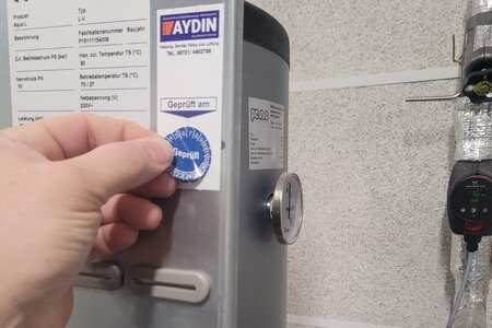 Ein blauer Prüfsticker mit der Aufschrift "Geprüft" wird von einer Hand auf einen silbernen Kasten geklebt.