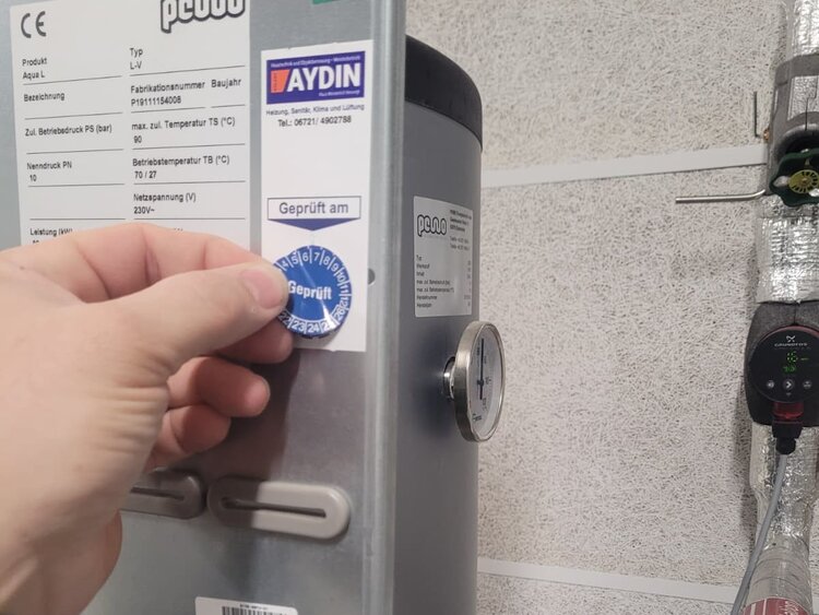 Ein blauer Prüfsticker mit der Aufschrift "Geprüft" wird von einer Hand auf einen silbernen Kasten geklebt.